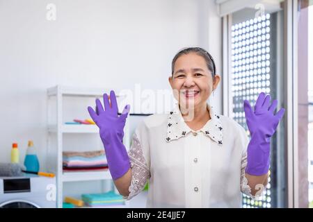 Asian senior elder woman wearing rubber gloves and rise hands in laundry room with proud, looking at camera, spring cleaning concept Stock Photo