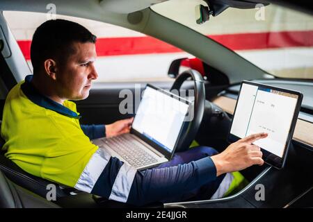 Male programmer with laptop working on digital tablet in electric car Stock Photo