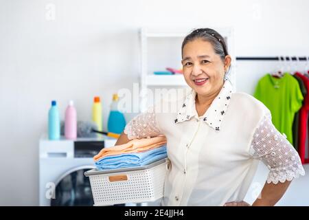 Happy smiling Asian senior elderly female woman carrying clothe basket doing laundry with washing machine, house cleaning and housekeeping, looking at Stock Photo