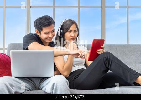Young Asian couple relaxing sitting on sofa using laptop computer and digital tablet working from home Stock Photo