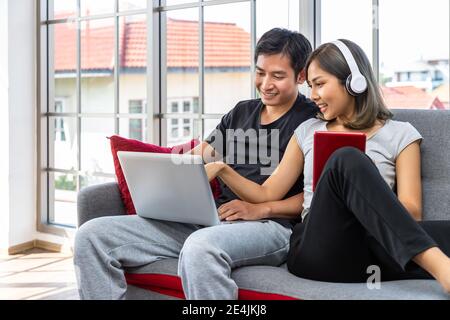 Young Asian couple relaxing sitting on sofa using laptop computer and digital tablet working from home Stock Photo