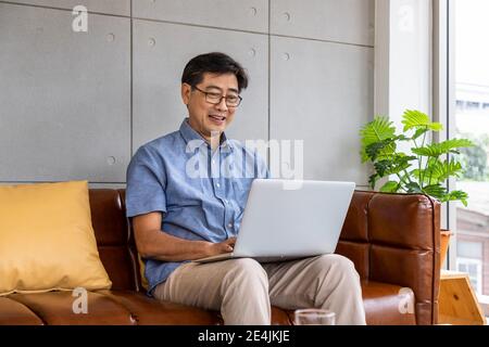 Happy Asian senior retired man sitting on couch sofa, using laptop computer Stock Photo