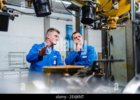 Male technicians wearing protective eyewear discussing on machinery equipment in factory Stock Photo