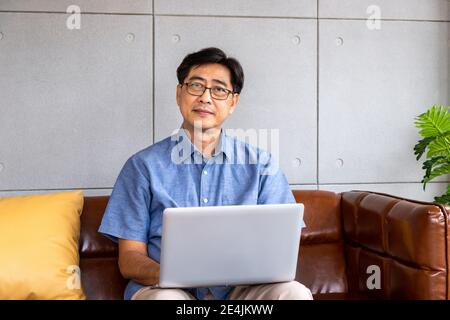Happy Asian senior retired man sitting on couch sofa, using laptop computer Stock Photo