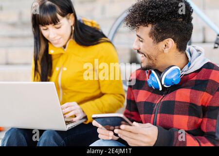 Man with digital tablet smiling while sitting by friend using laptop outdoors Stock Photo