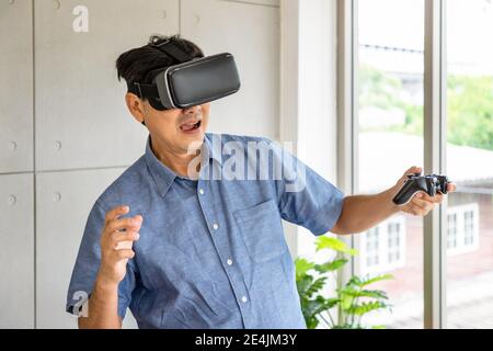 Senior retirement Asian man wearing virtual reality glasses standing holding joystick and playing game Stock Photo