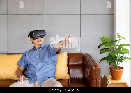 Senior retirement Asian man wearing virtual reality glasses sitting on couch enjoy exciting experience and pointing pushing button Stock Photo