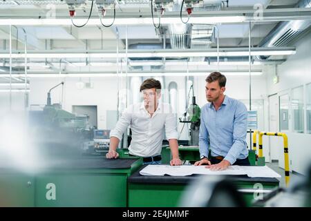 Male engineers with floor plan looking at manufacturing equipment while having meeting in factory Stock Photo