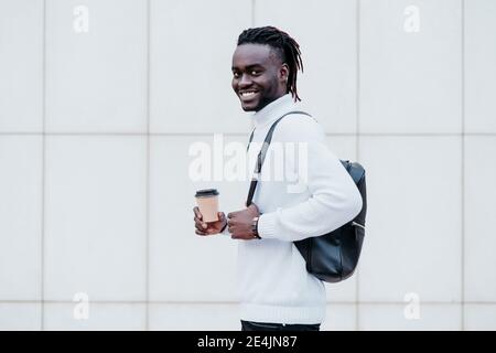 Young businessman carrying backpack while holding disposable coffee cup against white wall Stock Photo