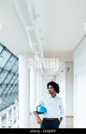 Young male engineer with hardhat looking away while standing in corridor at office Stock Photo