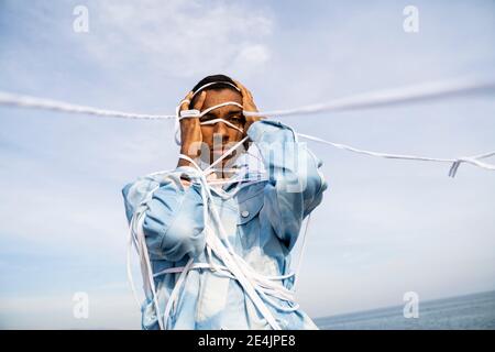 Man wrapped in rope standing with head in hands against sky Stock Photo