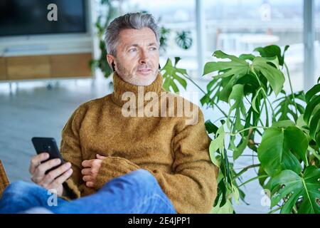 Mature man wearing in-ear headphones using mobile phone while sitting at home Stock Photo
