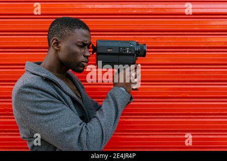 Man looking using 1980s film camera while standing against red wall Stock Photo
