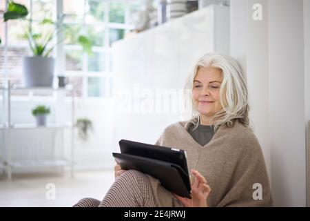 Smiling woman with white hair using digital tablet while sitting in living room at apartment Stock Photo