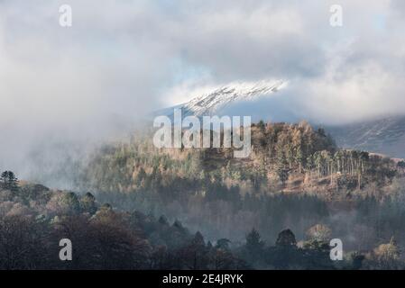 Epic landscape image looking across Derwentwater in Lake District towards Catbells snowcapped mountain with thick fog rolling through valley Stock Photo
