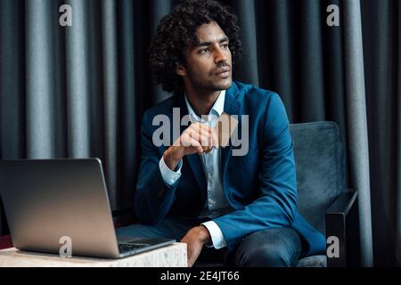 Thoughtful young male professional sitting with laptop holding credit card while looking away at office Stock Photo