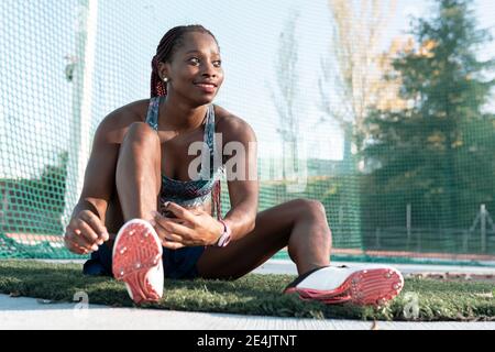 Smiling young sportswoman looking away while tying shoelace against net Stock Photo