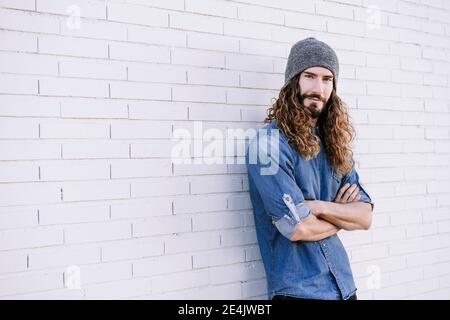 Young man wearing knit hat wit arms crossed leaning on brick wall Stock Photo