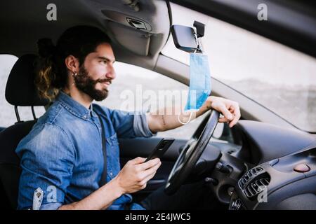 Smiling young man sitting in car while holding smart phone during COVID-19 Stock Photo