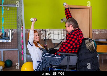 Young female nurse teaching disabled man to exercise with dumbbell at nursing home Stock Photo