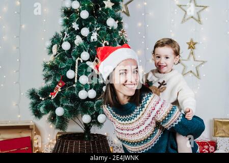 Smiling mother wearing Santa hat piggybacking cute daughter at home during Christmas Stock Photo