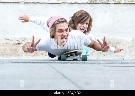 Smiling friends gesturing peace sign while lying on skateboard against footpath Stock Photo