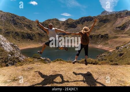 Couple holding hands while jumping over mountain on sunny day Stock Photo