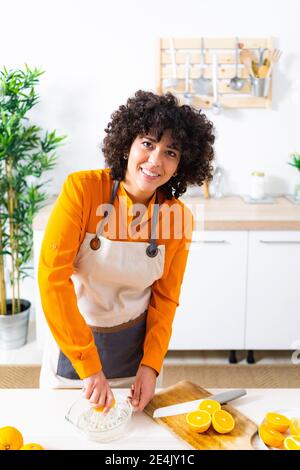 Young woman wearing apron squeezing orange for preparing juice while standing at home Stock Photo