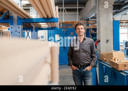 Portrait of carpenter posing with hands in pockets in front of shelf filled with wooden poles Stock Photo