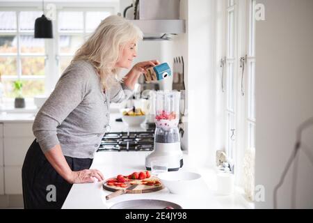 Senior woman pouring milk in processor while preparing strawberry smoothie at kitchen Stock Photo