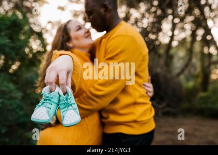 Pregnant woman holding baby shoe while embracing man standing at forest Stock Photo