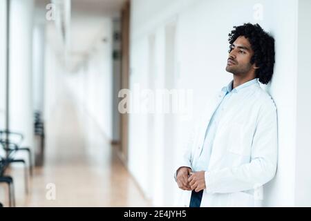 Thoughtful young male doctor leaning on white wall at hospital corridor Stock Photo