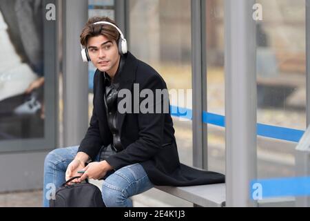 Handsome man listening music looking away while sitting at bus stop Stock Photo