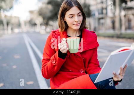 Young woman wearing winter jacket having coffee while reading book on street Stock Photo
