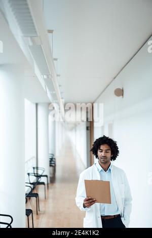 Confident young male doctor looking at clipboard in hospital corridor Stock Photo