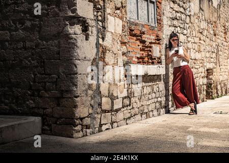 Woman leaning against old stone building wall and looking at smart phone Stock Photo