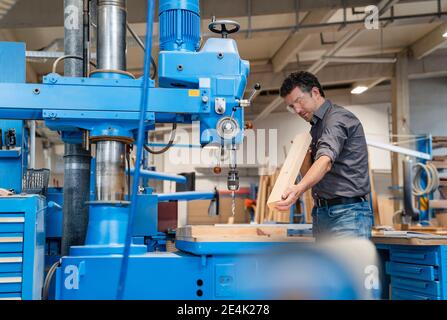 Carpenter wearing protective goggles drilling wooden plank in production hall Stock Photo