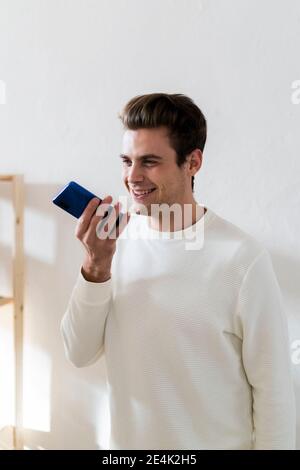 Smiling young man on speaker phone call against white wall at home Stock Photo