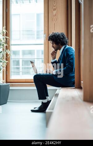 Young businessman using laptop while sitting against window at workplace Stock Photo