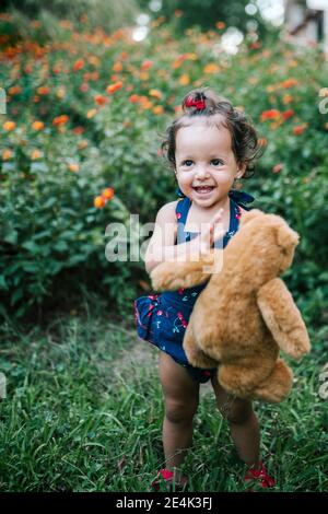 Cheerful baby girl holding teddy bear standing on grassy land against plants in park Stock Photo