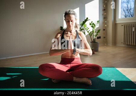 Mature woman with hands clasped doing yoga on exercise mat in sunlight at home