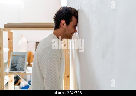 Smiling young man with eyes closed banging head on white wall in living room