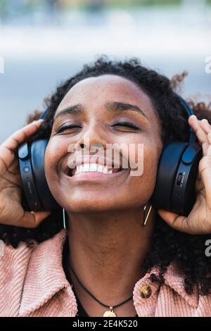 Happy afro woman with eyes closed listening music through headphones in city