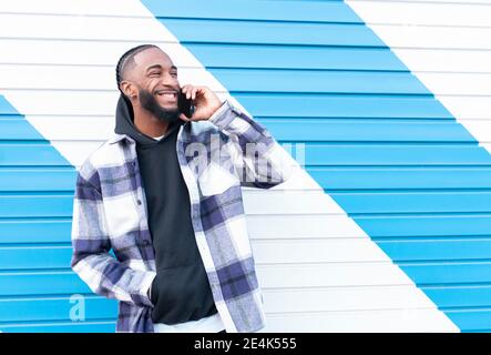 Happy young man with hands in pockets on phone call looking away against corrugated iron Stock Photo