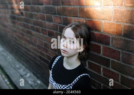 Serious teenage girl with blue eyes leaning on brick wall in city Stock Photo