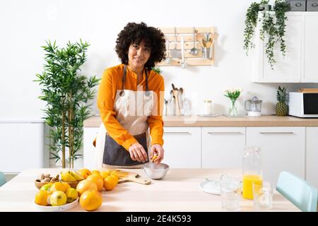 Smiling woman squeezing orange in bowl while standing at kitchen Stock Photo