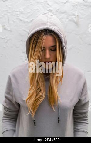 Close-up of sad woman with eyes closed wearing hood while standing against white wall Stock Photo