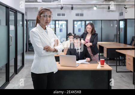 Young asian businesswoman confident with crossed arms wearing face shield and coworkers seriously working on desk at new normal office Stock Photo