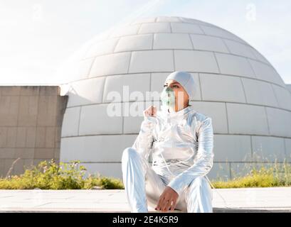 Young woman wearing protective suit and oxygen mask standing with hand raised against igloo Stock Photo