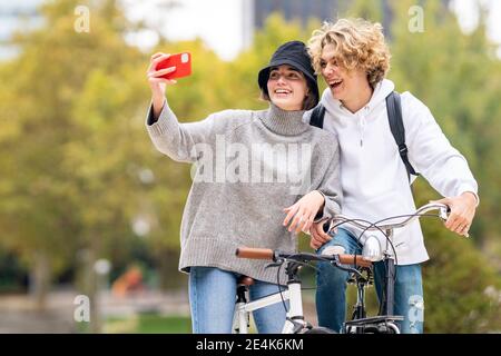 Happy woman taking selfie standing by male friend with bicycle in park Stock Photo